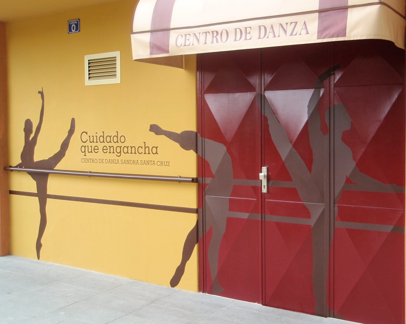 Fachada pintada a tamaño real con la silueta de tres bailarines en diferentes poses y en medio de dos se lee "Cuidado que enganca" y debajo, El Centro de Danza Sandra Santa Cruz permanecerá cerrado y continuará con sus clases on line hasta que sea seguro el regreso a clase.