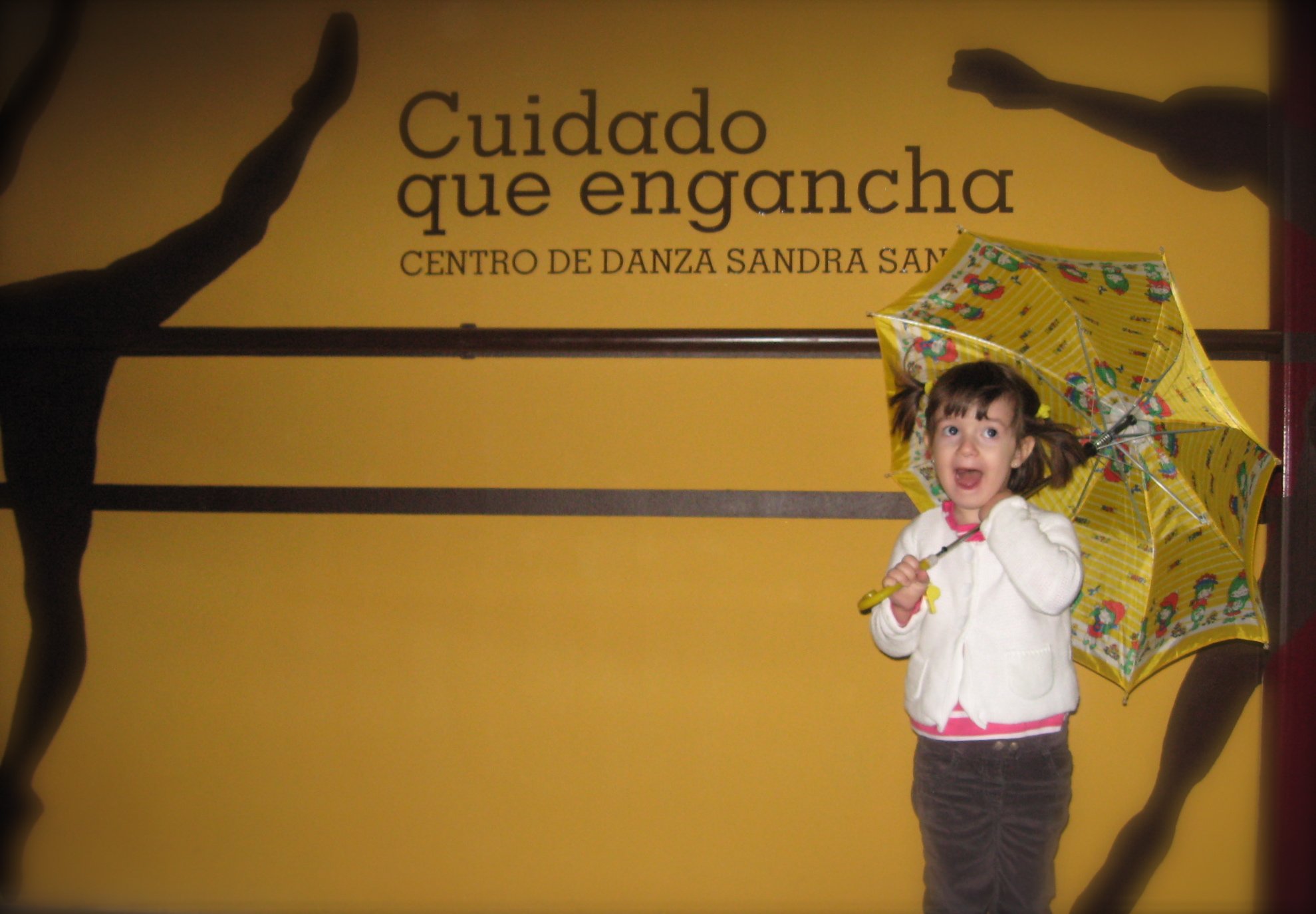 Foto divertida de una alumna de 5 años con dos coletas y la boca abierta en la fachada del Centro de Danza Sandra Santa Cruz agarrada a su paraguas abierto que apoya sobre un hombro.