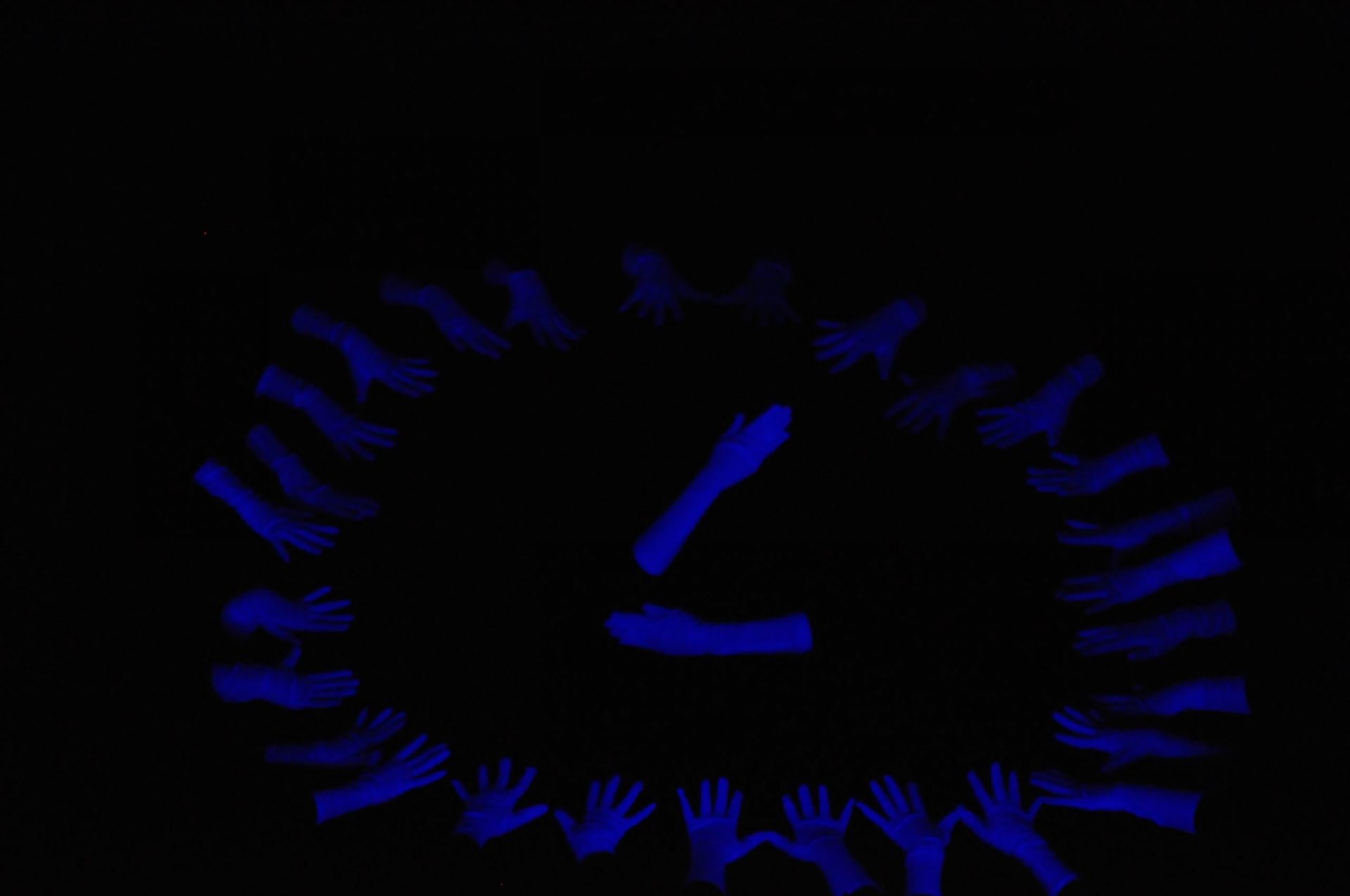 Foto de la coreografía de luz negra "Manos" del Centro de Danza Sandra Santa Cruz que bailará en el Día Mundial de la Danza. Un grupo de manos  con guantes blancos hasta el codo forman el círculo de un reloj y en el centro, otras manos hacen de minutero y segundero. 