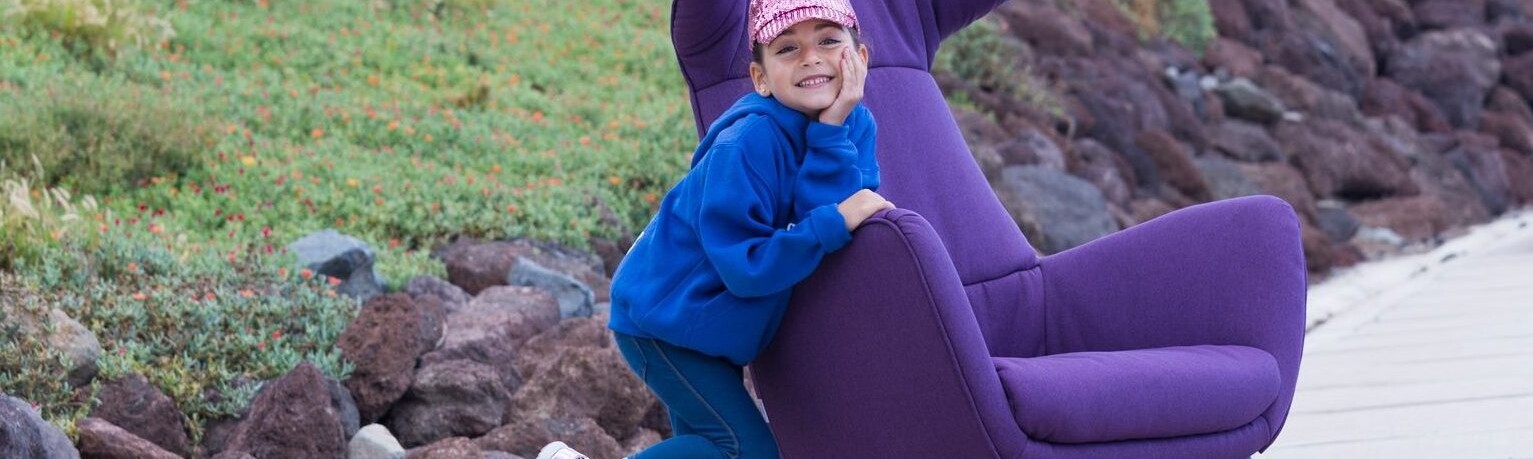 Foto de una alumna del Centro de Danza Sandra Santa Cruz de 6 años en el paseo de la playa de La Laja. Lleva un vaquero largo azul con una sudadera azulina y una visera sora plastel de lentejuelas monísima a juego con sus playeras que son exactamente igual que la gorrita. Está apoyada en el brazo de un enorme sillón orejero malva de manera divertida con una enorme sonrisa. Tiene una en la carita y un pie levantado-Fotografía Ethel Bartrán