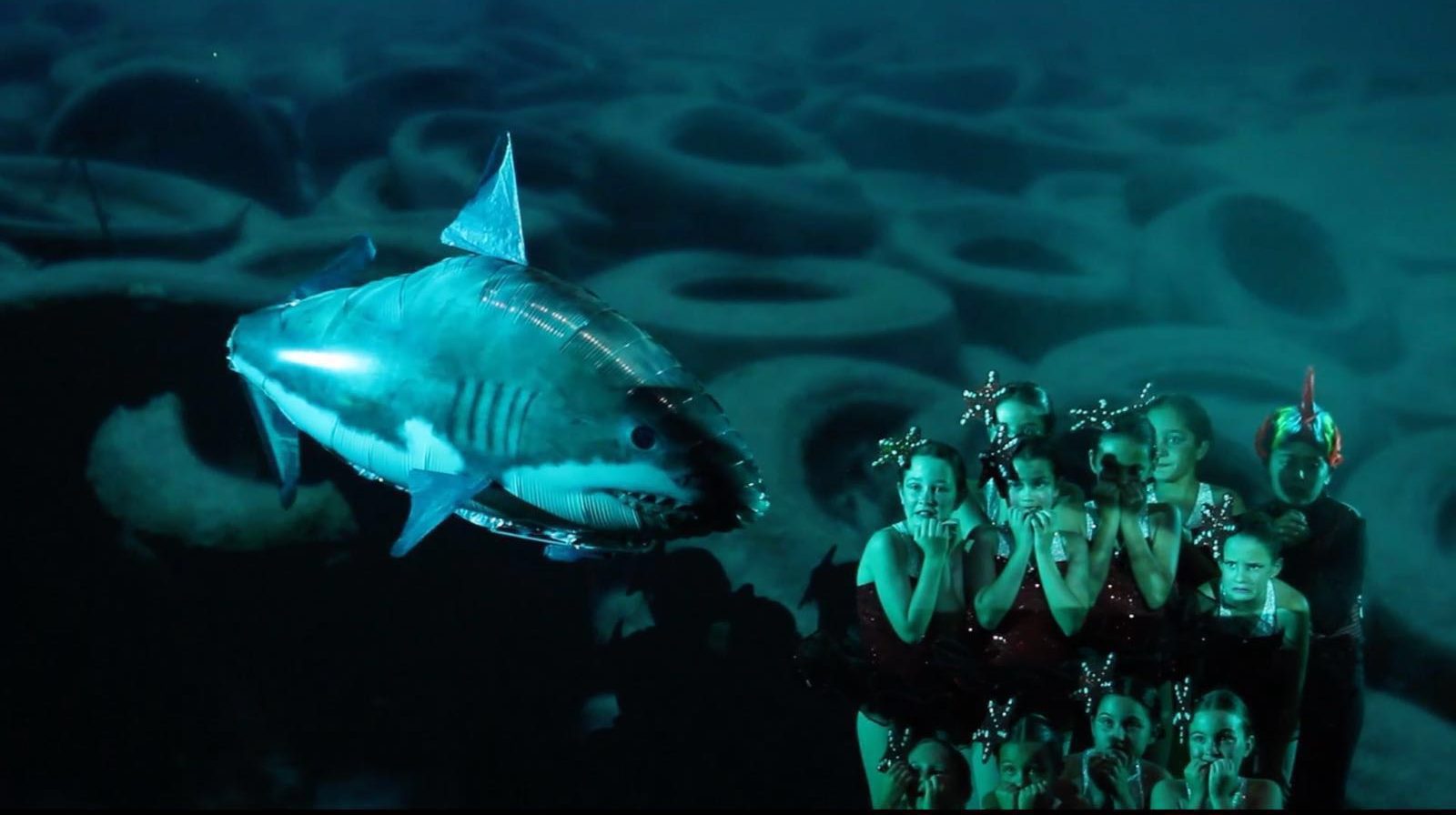 Si los peces hablaran... Musical del Centro de Danza Sandra Santa Cruz. Foto del Musical Si los peces hablaran... del Centro de Danza Sandra Santa Cruz donde un tiburón de helio se acerca amenazante a un grupo baialrines vestidos de peces que lo mira aterrado. En la pantalla de proyección se ve que el fondo marino está lleno de gomas de neumáticos que se han arrojado al mar.