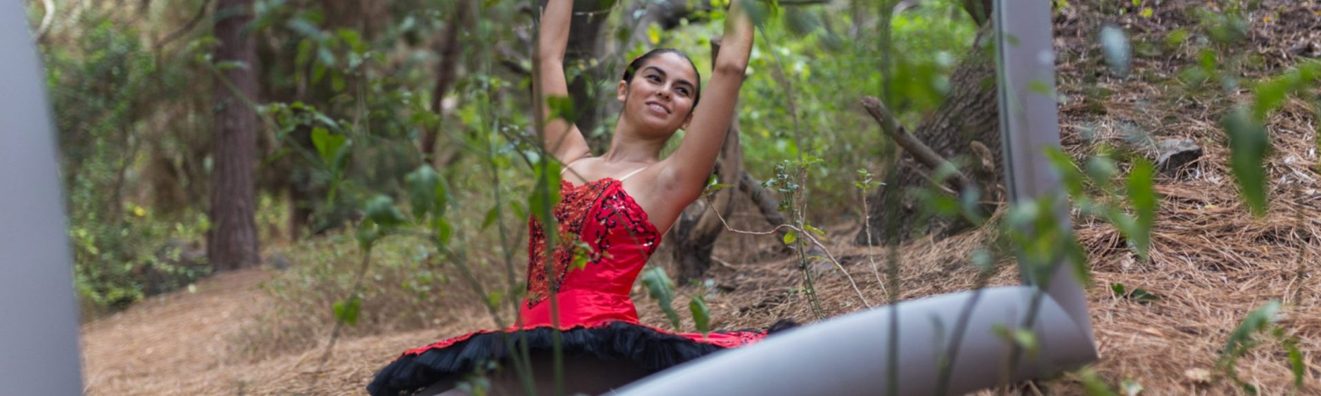 Foto de una bailarina del Centro de Danza Sandra Santa Cruz en el campo sentada con los pies estirados y haciendo un cambré ante un gran espejo. Tiene zapatillas de puntas y un espectacular tutú de plato en color rojo con adornos en negro, para explicar que el trabajo en el Centro es: Muchomasquedanza