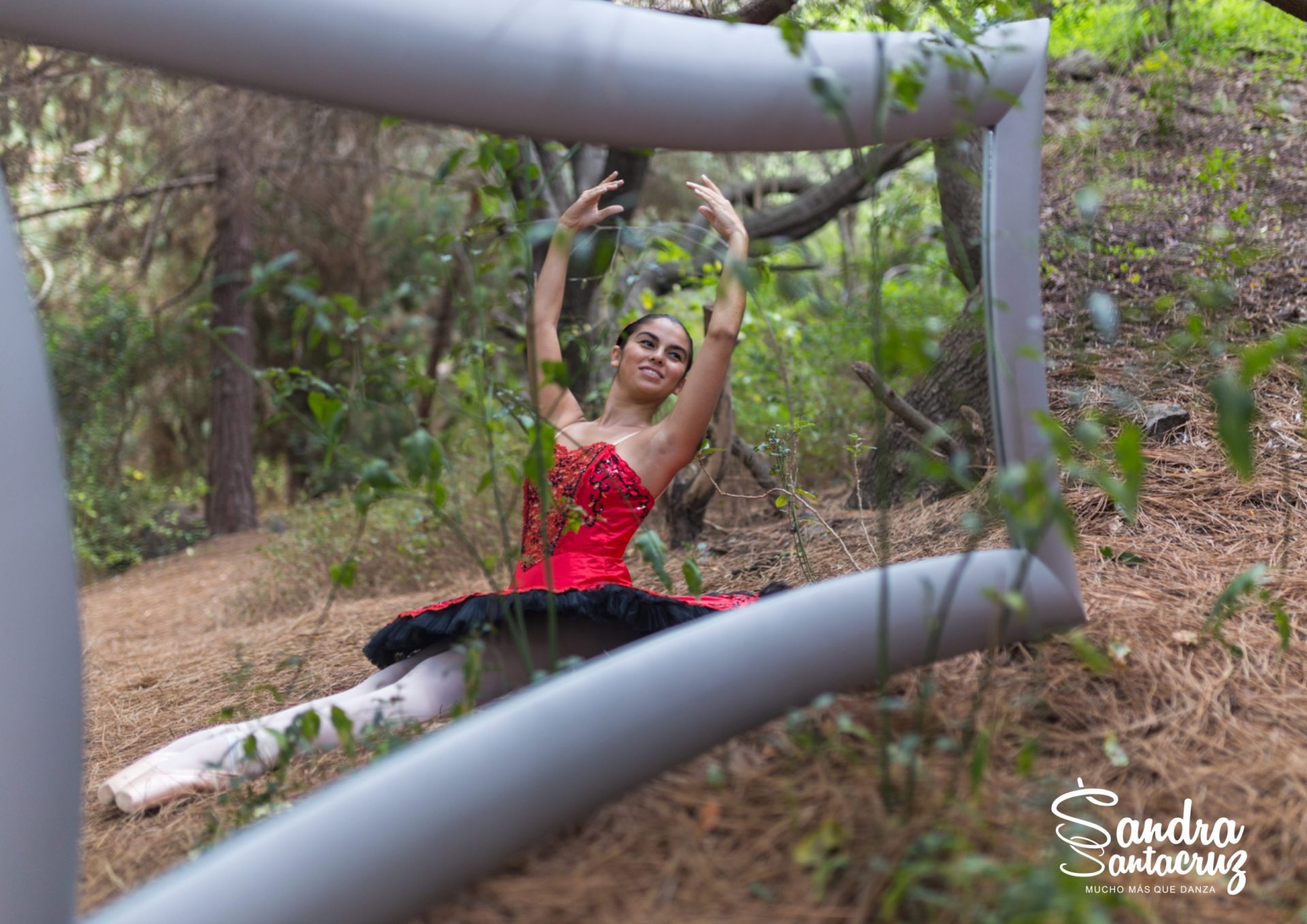Foto de una bailarina del Centro de Danza Sandra Santa Cruz en el campo sentada con los pies estirados y haciendo un cambré ante un gran espejo. Tiene zapatillas de puntas y un espectacular tutú de plato en color rojo con adornos en negro, para explicar que el trabajo en el Centro es: Muchomasquedanza. Fotografía Ethel Bartrán.