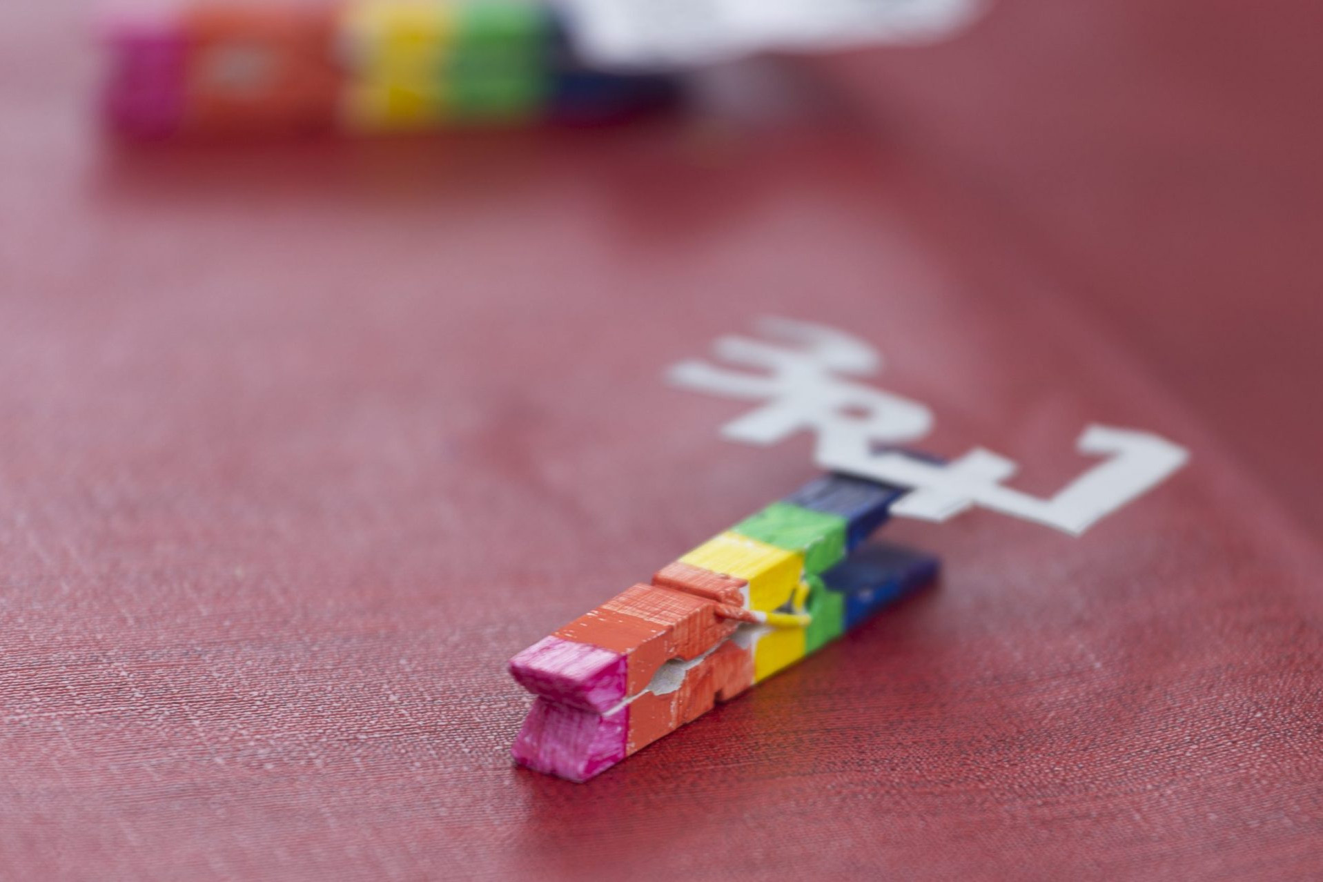 Foto de una traba de la ropa pintada con los colores del arcoíris y lleva pegado en material recicado con brick de leche la frase 3R+1. Taller de reutilización de Sandra Santa Cruz.