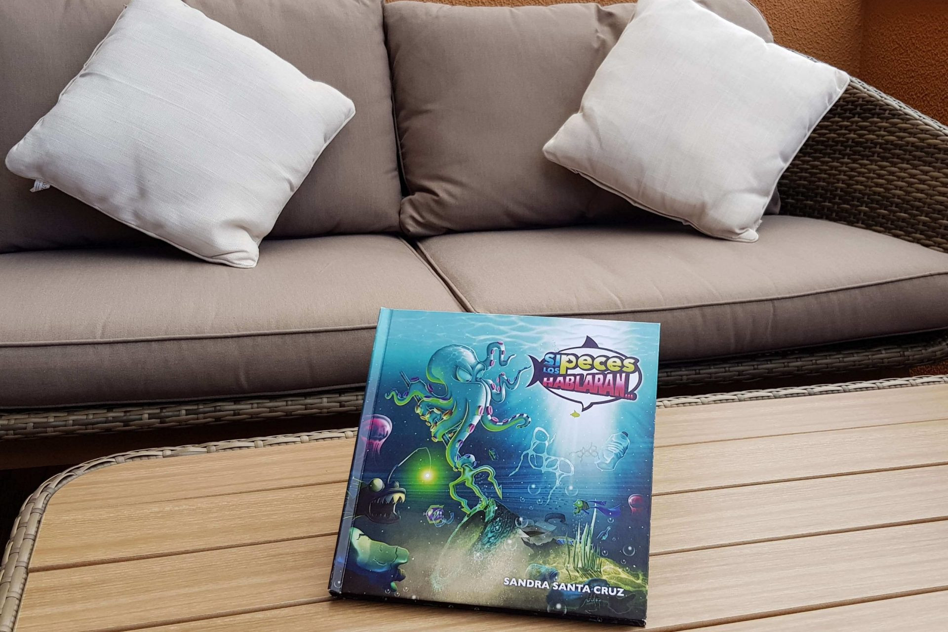Foto del cuento Si los peces hablaran... de Sandra Santa Cruz sobre una mesa preparado para la entrevista con Radio Dunas. Detrás un cómodo sofá con cojines invitan a la charla.