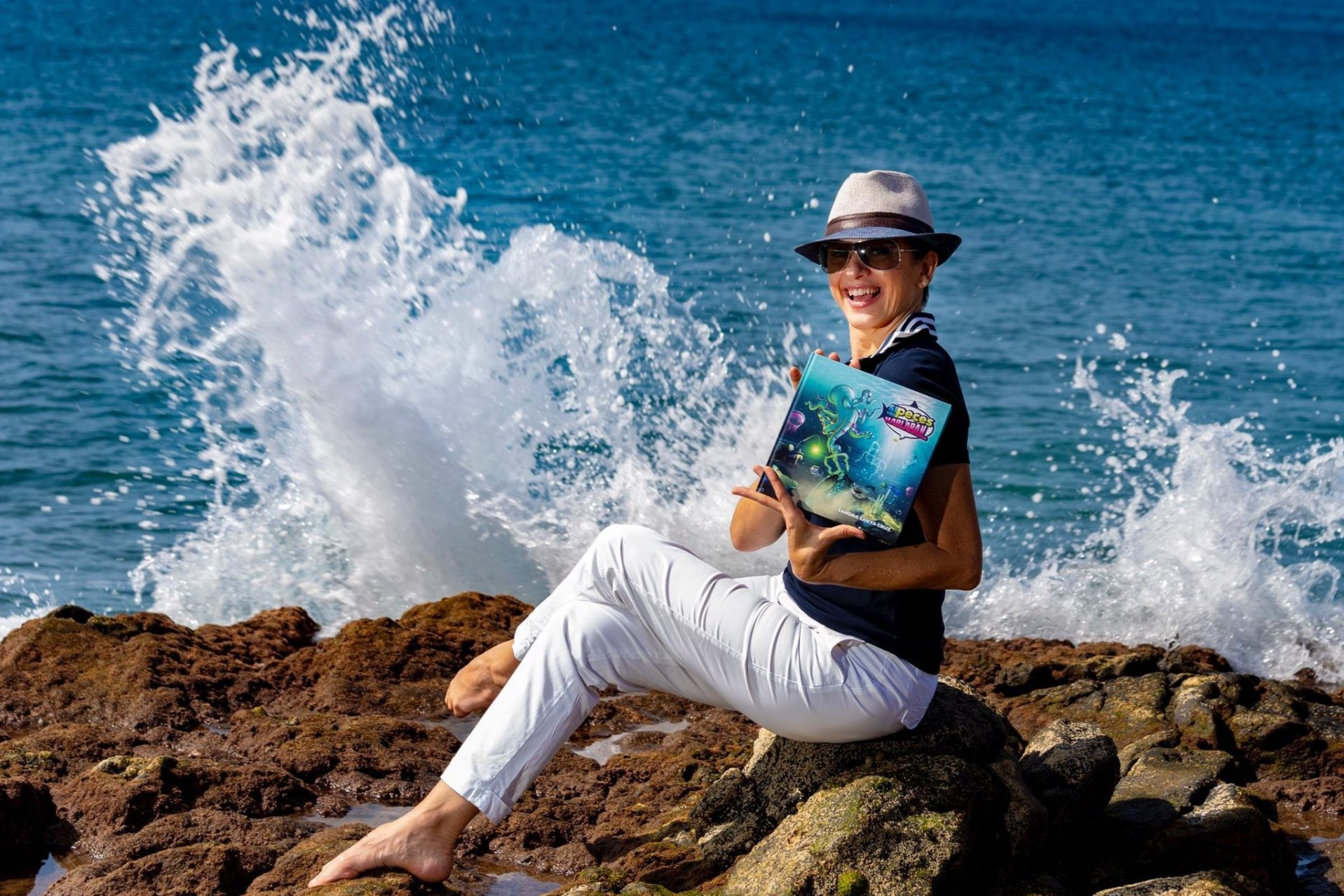 Foto de Sandra Santa Cruz sentada en unas rocas con su cuento Si los peces hablaran... en las manos, con el mar de fondo y unas olas rompiendo detrás de ella. Su cuetno quiere ayudar a proteger el mundo marino y frenar la contaminación. Entrevista en la revista Enjoy