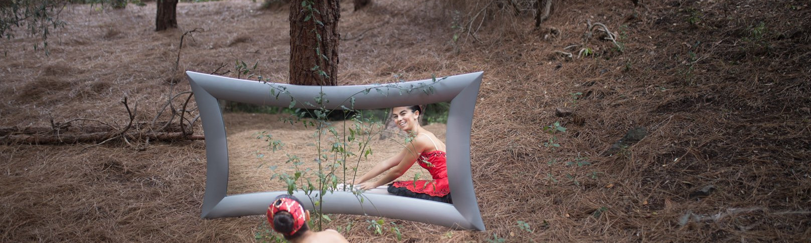 Foto de una bailarina del Centro de Danza Sandra Santa Cruz en el campo sentada con los pies estirados y las manos apoyadas en los pies mirándose sonriéndo ante un gran espejo. Tiene zapatillas de puntas y un espectacular tutú de plato en color rojo con adornos en negro, para explicar que el trabajo en el Centro es: Muchomasquedanza