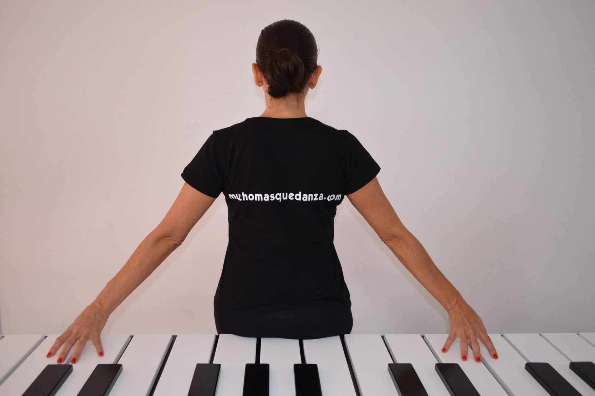 Foto de la directora del Centro de Danza Sandra Santa Cruz de espaldas sentada sobre las teclas de un piano gigantesco y en cuya camiseta se lee la filosofía del Centro: Muchomasquedanza.