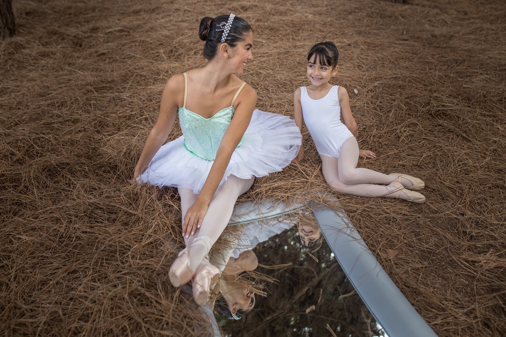 Foto de dos bailarinas del Centro de Danza Sandra Santa Cruz en el campo, sentadas sobre pinocha que tienen junto a ellas un enorme espejo. La adolescente, que lleva un tutú de plato verde pastel, zapatillas de puntas, moño y diadema de perlas, tiene una mano en la pinocha y la otra sobre sus pies, que están cruzados sobre el espejo, en donde se refleja un poco de sus puntas, el tul de su tutú y su cara sonriente mirando a la más pequeñita, que lleva puesto simplemente una malla blanca y moño. Con los pies recogidos fuera del espejo y a poyada hacia atrás sobre sus manos, solo se ve su bonita sonrisa reflejada en el espejo. Irradia complicidad además de armonía. Lo que pretende que descubras nuestro Centro con la clase de Slim Ballet para adultos, que te proponeos para ponerte en forma.