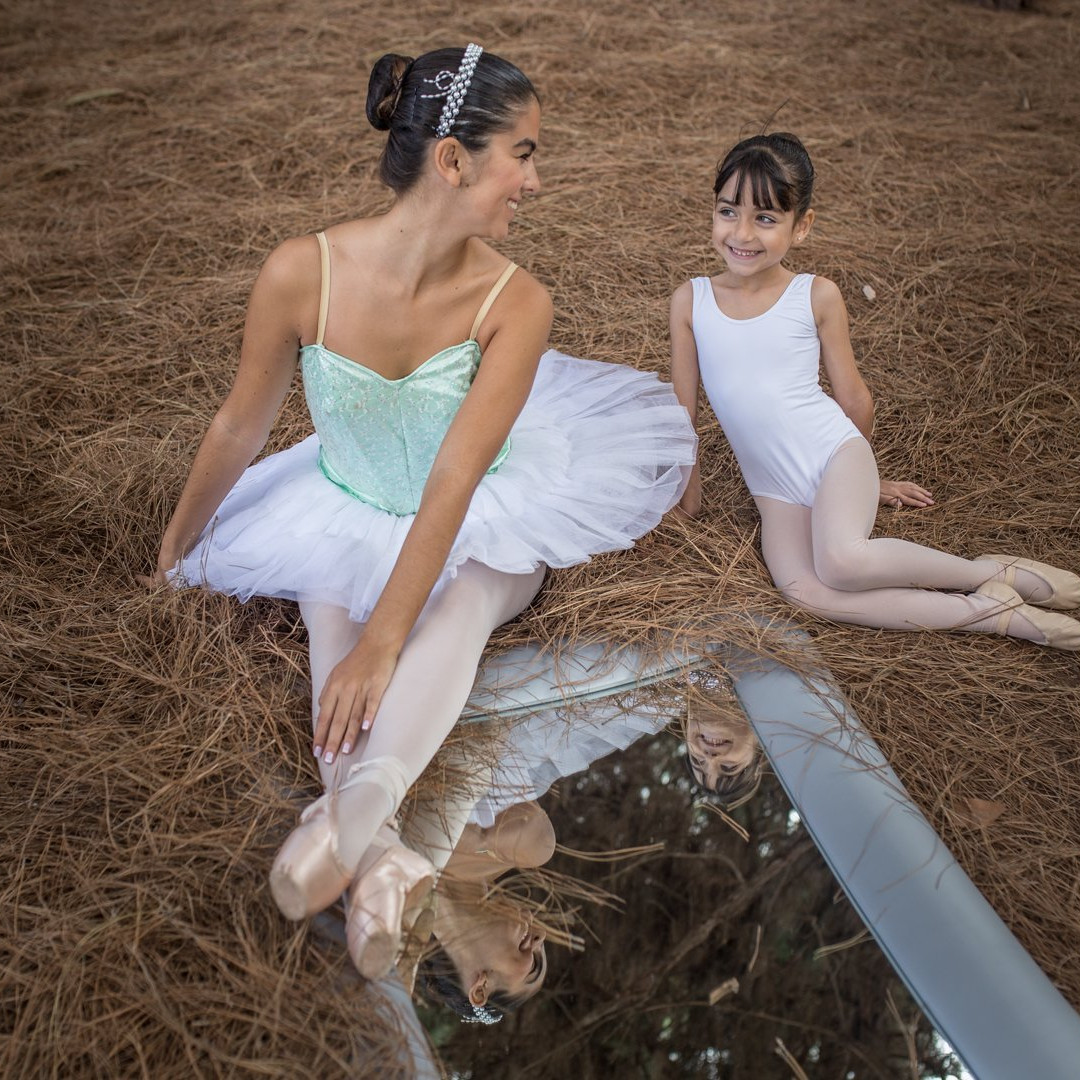 Foto de dos bailarinas del Centro de Danza Sandra Santa Cruz en el campo, sentadas sobre pinocha que tienen junto a ellas un enorme espejo. La adolescente, que lleva un tutú de plato verde pastel, zapatillas de puntas, moño y diadema de perlas, tiene una mano en la pinocha y la otra sobre sus pies, que están cruzados sobre el espejo, en donde se refleja un poco de sus puntas, el tul de su tutú y su cara sonriente mirando a la más pequeñita, que lleva puesto simplemente una malla blanca y moño. Con los pies recogidos fuera del espejo y a poyada hacia atrás sobre sus manos, solo se ve su bonita sonrisa reflejada en el espejo. Irradia complicidad además de armonía. Lo que pretende que descubras nuestro Centro con la clase de Slim Ballet para adultos, que te proponeos para ponerte en forma.