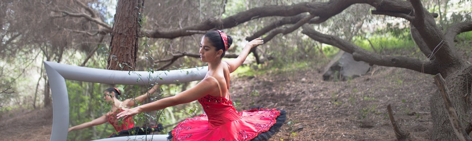 Foto de Olga Ortega, bailarina del Centro de Danza Sandra Santa Cruz en el campo vestida con un maravilloso tutú rojo de rodillas en 4ª posición y apoyada en una de sus zaaptillas de puntas con los brazos en 4ª alonyé, posando delante de un gran espejo espejo plateado de situeta con forma sinuosa
