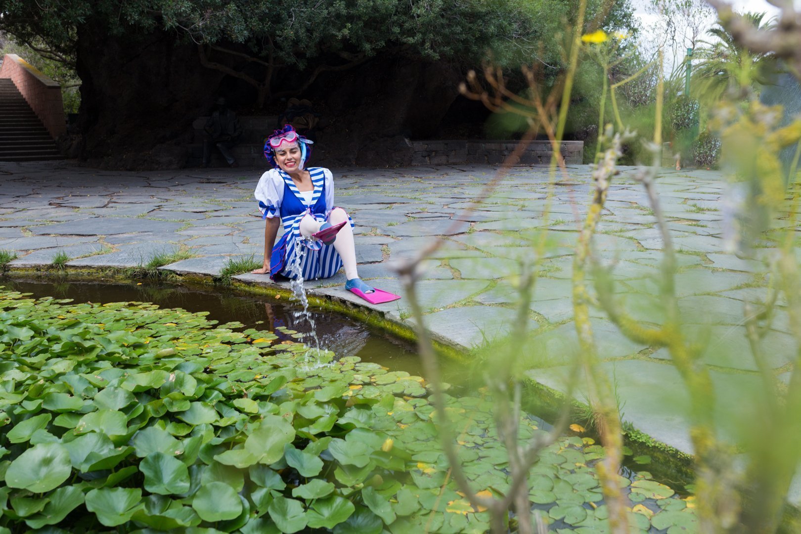 foto de una aluman del Centro de Danza Sandra Santa Cruz en el Jardín Canario con gafa, tubo, aletas y un vestuario de nadadora del siglo pasado slapicando el agua de una fuente con una de sus aletas