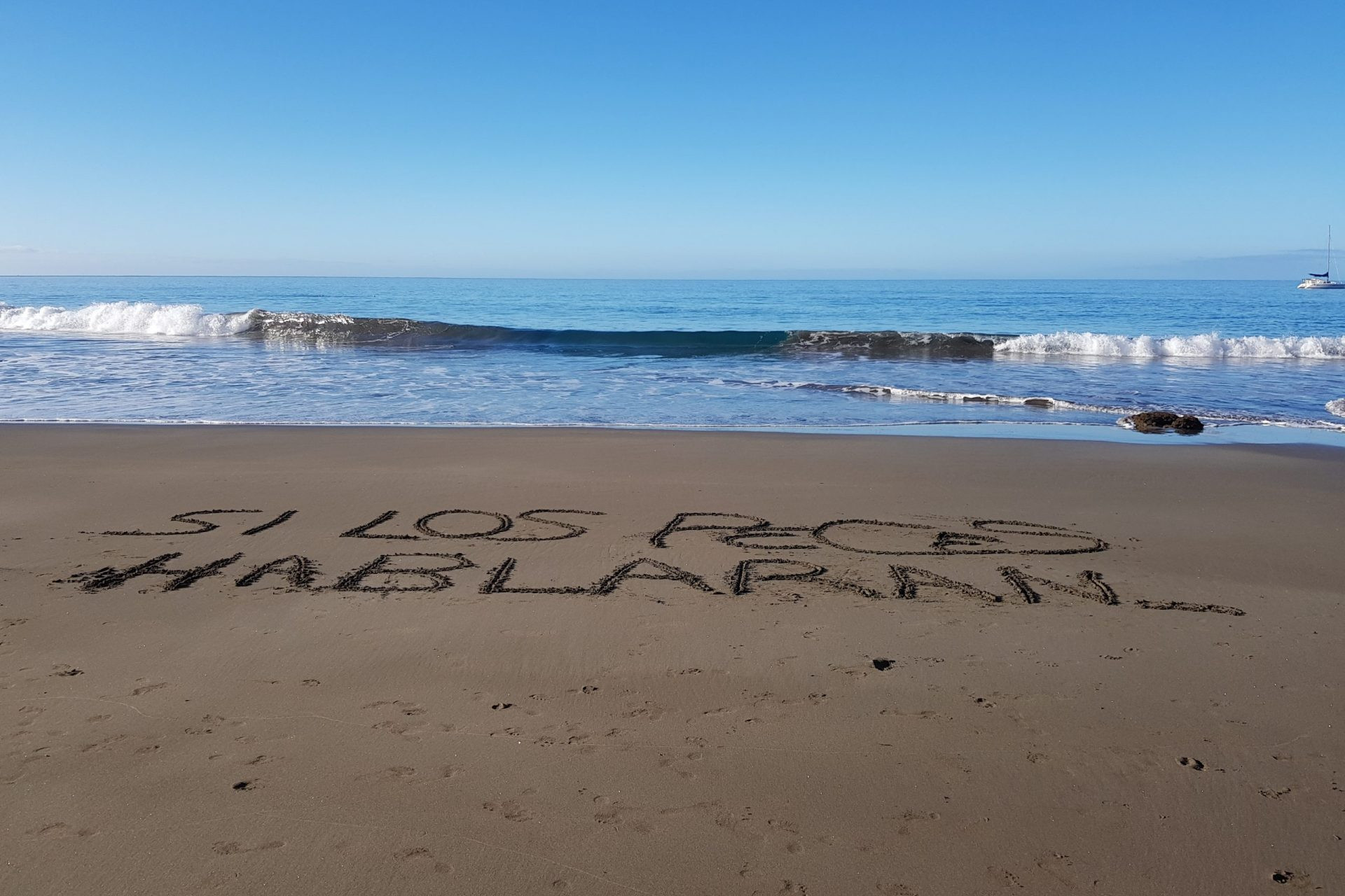Foto en la playa de Patalavaca de una ola acercándose a la orilla donde en la arena está escrito 
