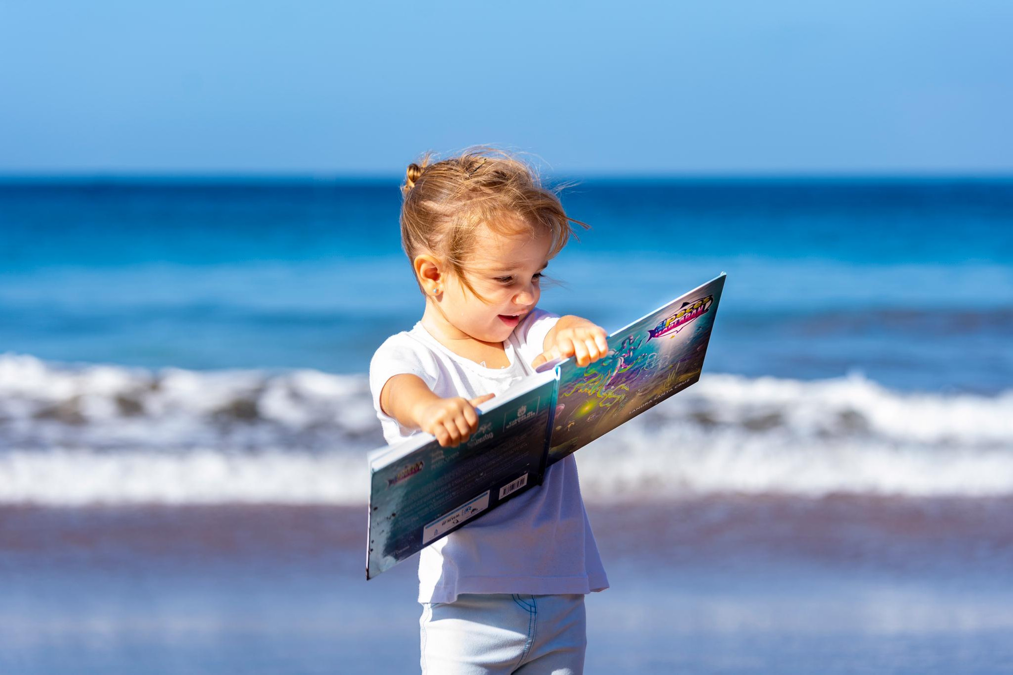 Foto de una niña pequeña en orilla de la playa leyendo el cuento Si los peces hablaran... con una enorme sonrisa.Cuento con valores de Sandra Santa Cruz.