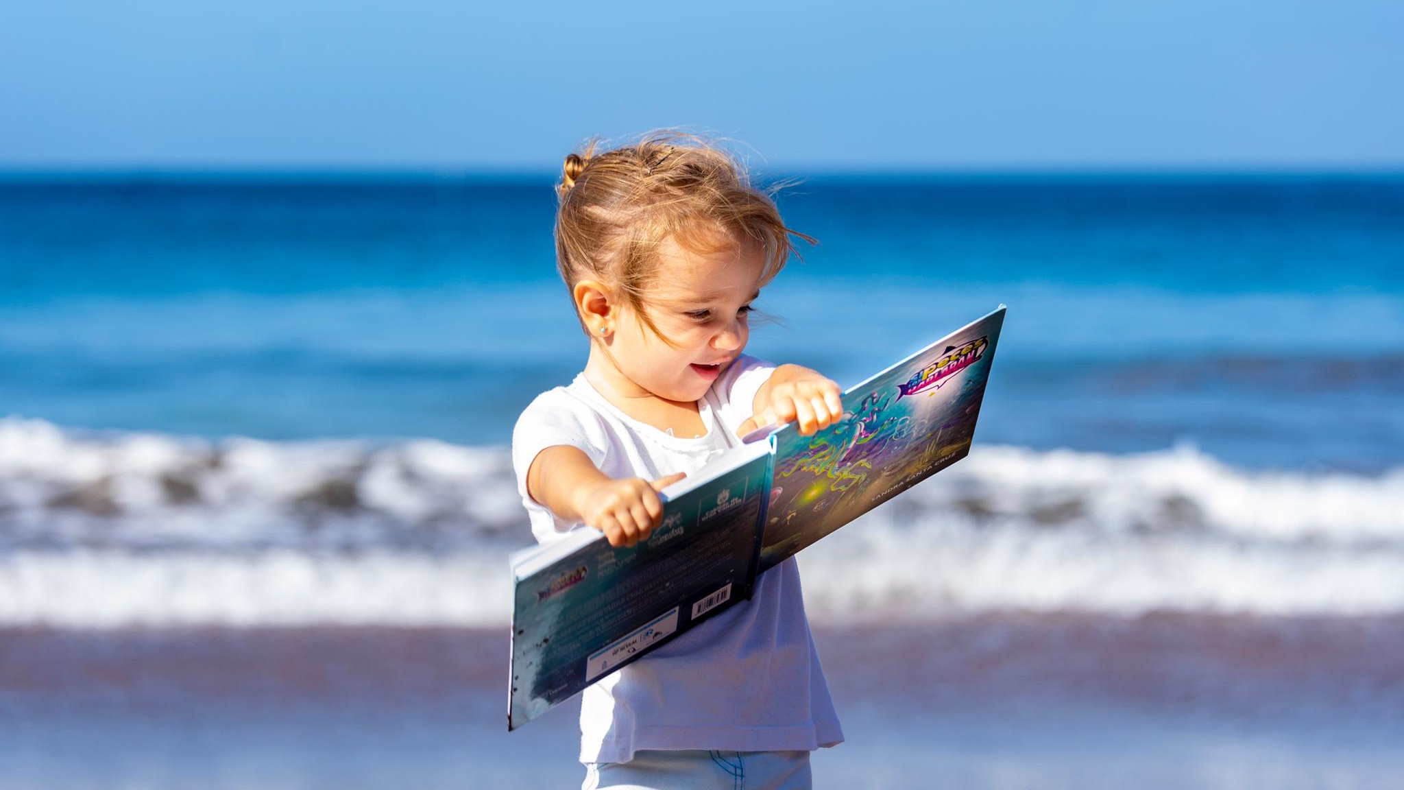 Foto de una niña pequeña en orilla de la playa leyendo el cuento Si los peces hablaran... con una enorme sonrisa.Cuento con valores de Sandra Santa Cruz.