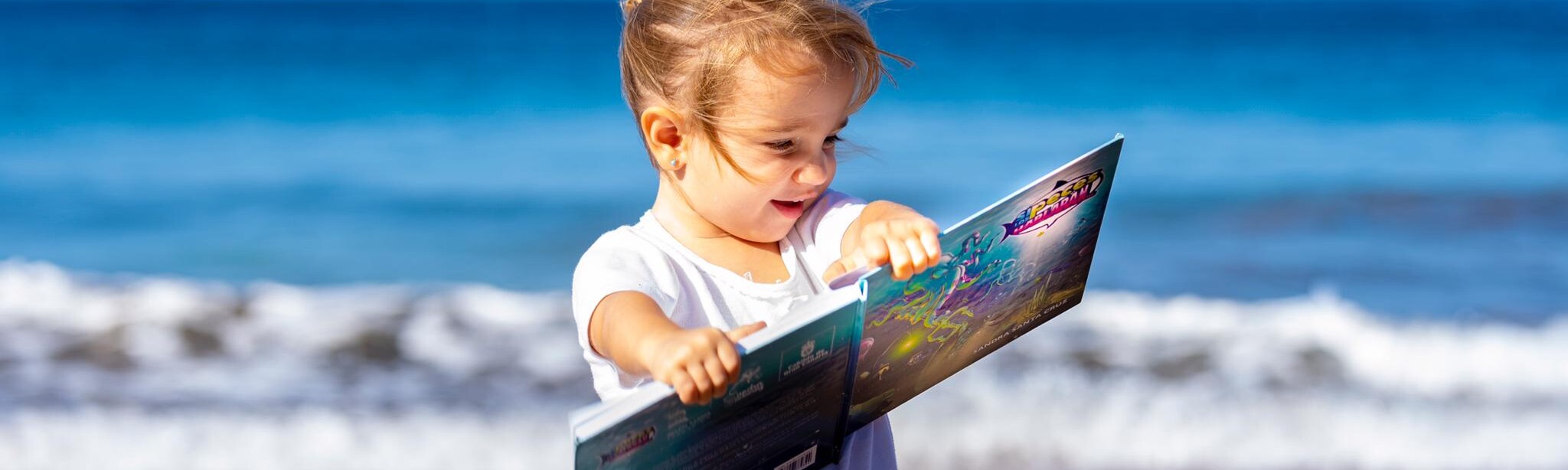 Foto de una niña pequeña en orilla de la playa leyendo el cuento Si los peces hablaran... con una enorme sonrisa.Cuento con valores de Sandra Santa Cruz.