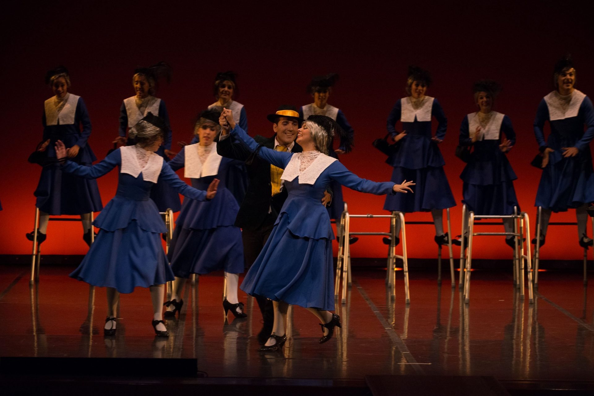 Foto de un fragmento del Musical: Los Productores donde hay grupo de 10 bailarines haciendo de ancianas. 7 están bailando subidas a sus taca-tacas y 3 con Mac Bially. Las bailarinas del Centro de Danza Sandra Sandra Santa Cruz van vestidas con un traje de dos pieza azul petróleo y un gran cuello de encaje blanco, ataviadas con tacones de anciana, eso sí,de purpurina negra, bosito de mano haciendo juego y por supuesto, pelucas blancas con moño bajo, que tiene incorporado un casquete negro con un velo corto de tul negro.