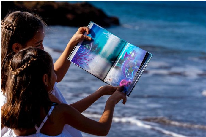 Foto de 2 niñas en la playa leyendo el cuento de Si los peces hablaran... con el mar de fondo, y donde apararece 