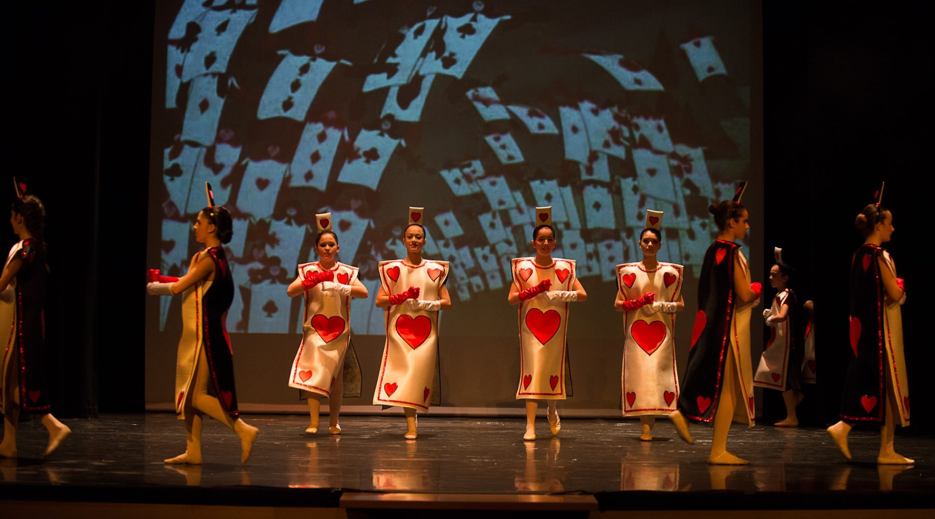 Foto del espectáculo "Con mis mejores deseos" del Centro de Danza Sandra Santa Cruz. En un fragmento de Alicia en el país de las maravillas donde un grupo considerable de bailarinas que interpretan el papel de las cartas hacen diferentes figuras como si se estuviesen barajando. En rigurosa formación la escena se refuerza con una diapositiva de la película que simboliza este momento.
