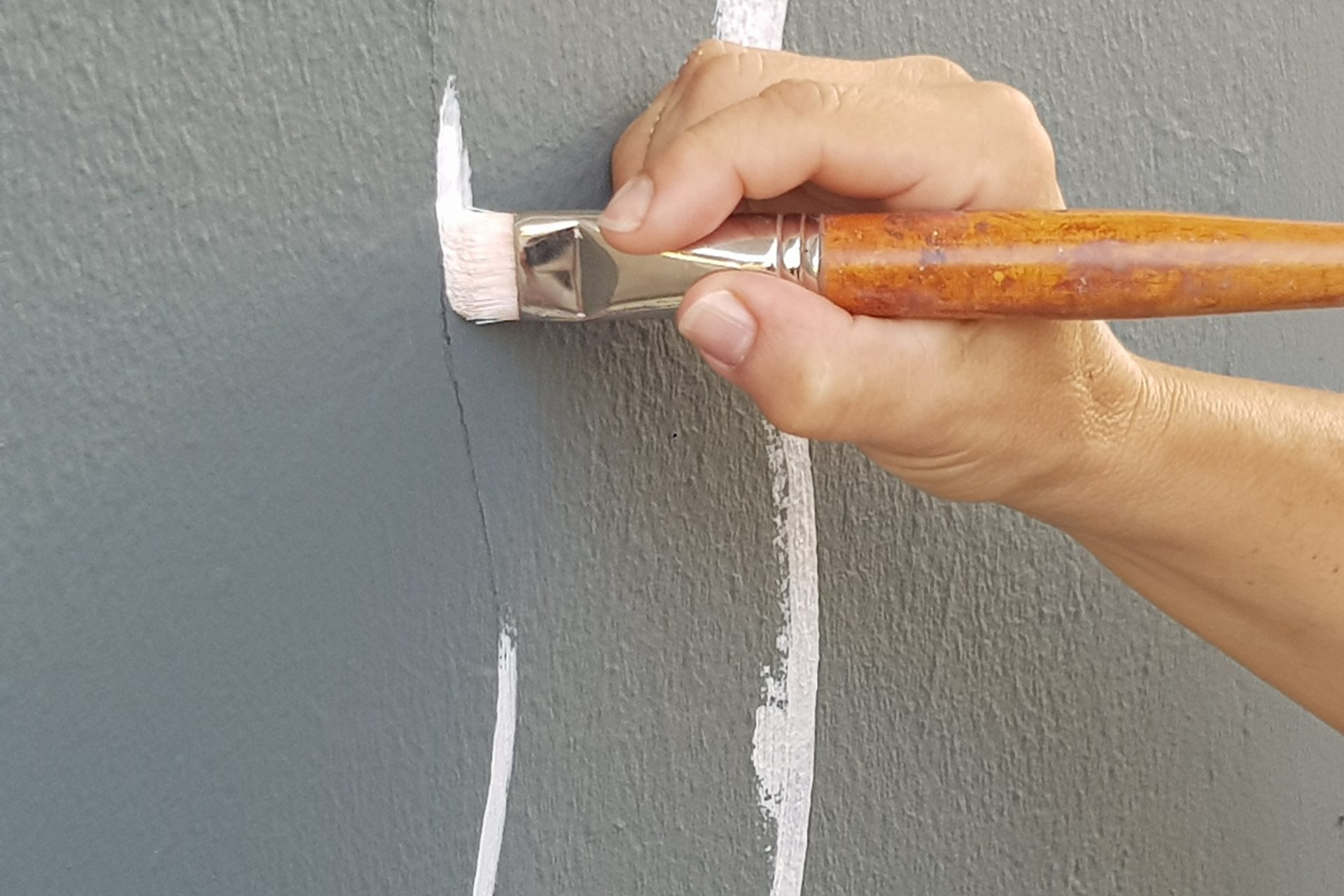 Foto de unas manos pintando en una fachada unos trazos largos en color blanco. Es la fachada del Centro de Danza Sandra Santa Cruz, que se prepara para el curso escolar 21/22 pintando a tamaño real, unos bailarines en su fachada. Las manos son de la pintora, Lidia Díaz.