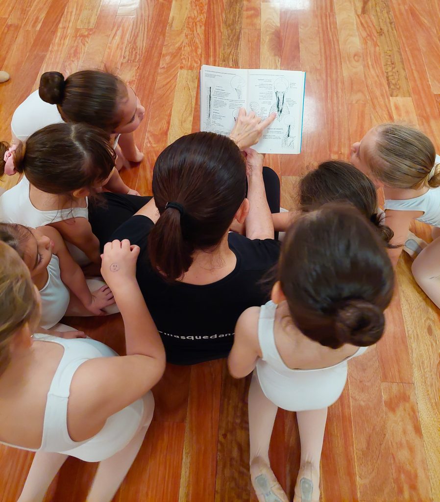 Foto de la directora del Centro de Danza Sandra Santa Cruz con un libro de anatomía en las manos y rodeada de su alumnado de Predanza explicándoles la importancia de la en danza. Anatomía y danza van de la mano en nuestro Centro.